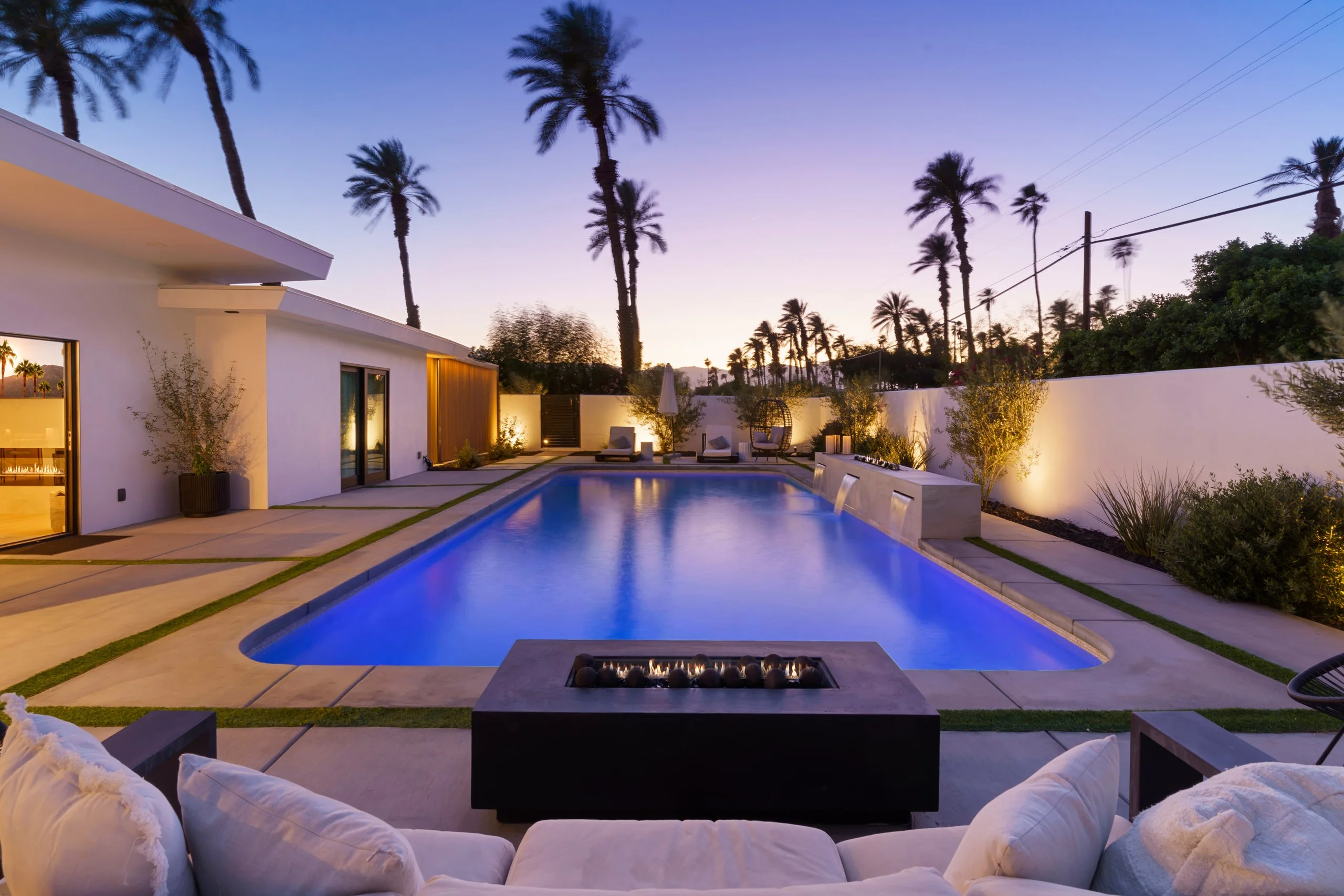 Long pool at dusk with a lit fire table in the foreground, palms silhouetted against a violet sky