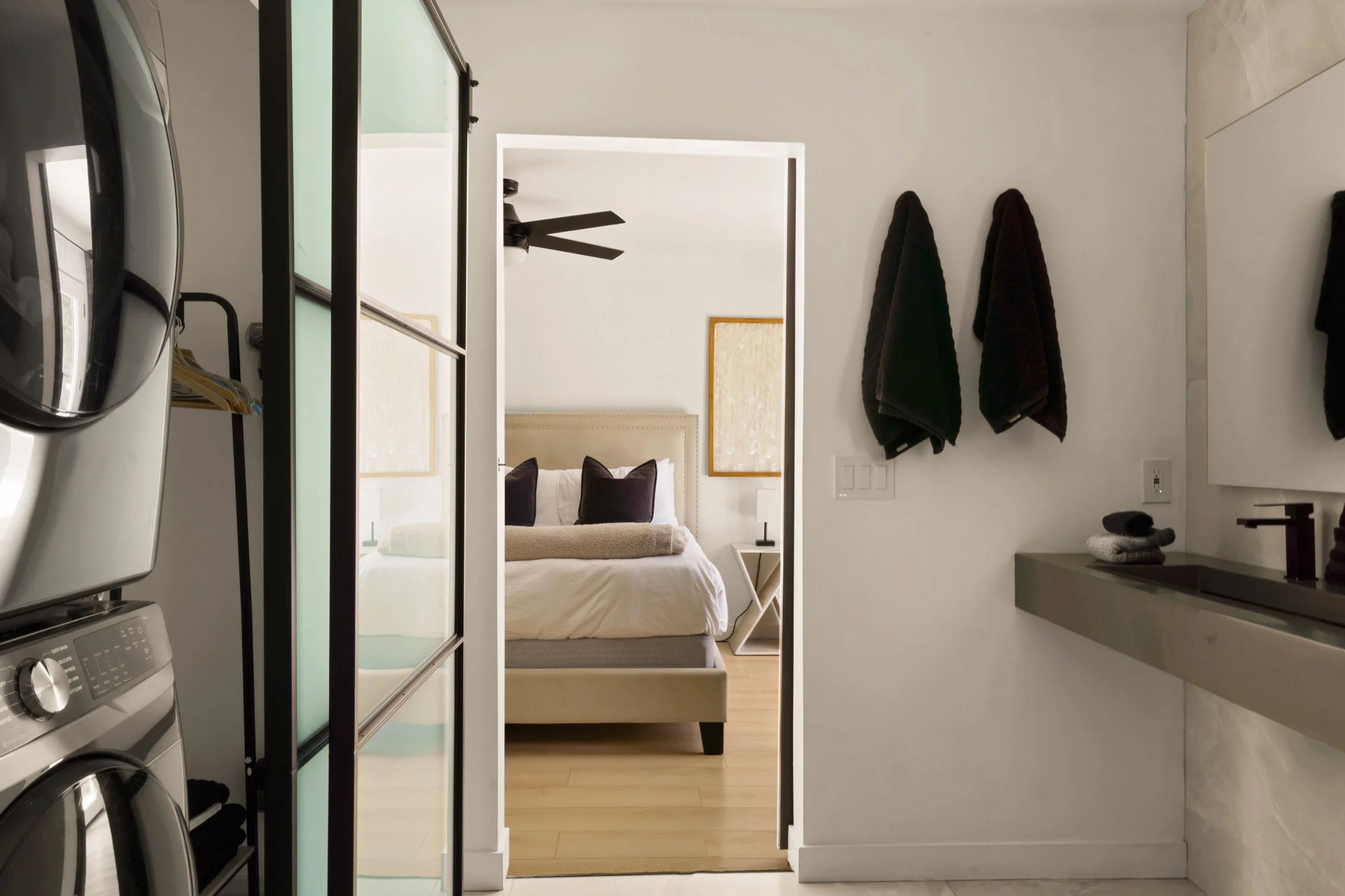 View through an open steel-framed door to a made bed, robes hanging on the wall beside a stone vanity