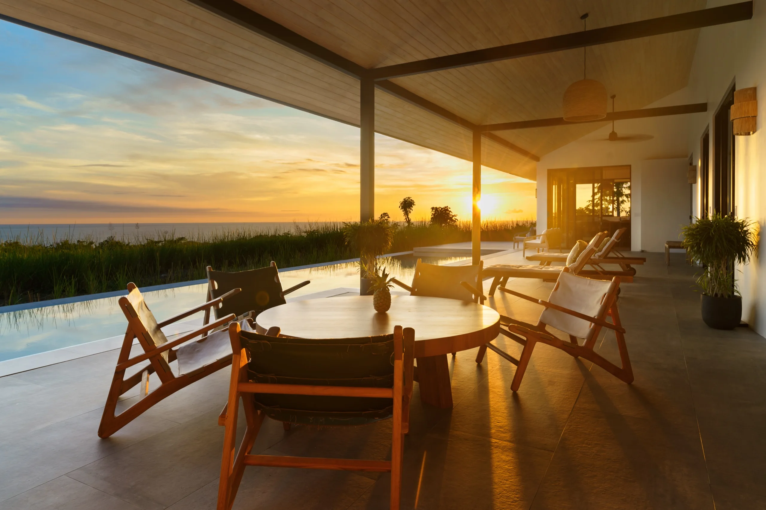 Covered terrace at golden hour, teak chairs around a round table, the pool edge and dune grass beyond