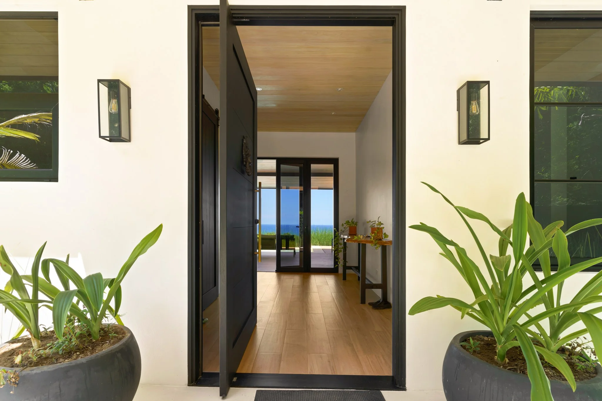 Entry with a black pivot door standing open, planted urns either side and the ocean framed straight through the house