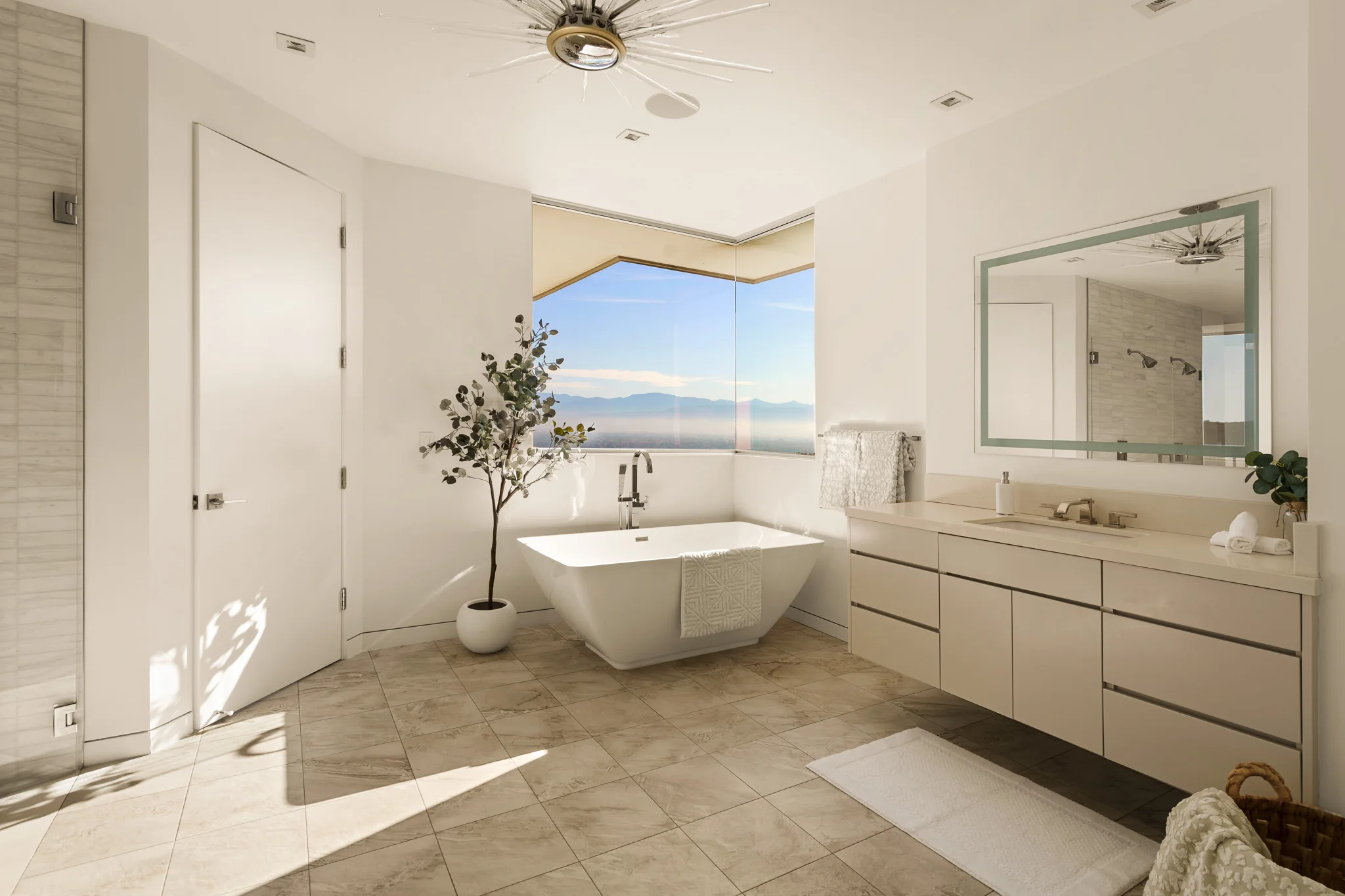 Freestanding tub in a white bathroom, olive tree in a stone pot and a corner window over the hills