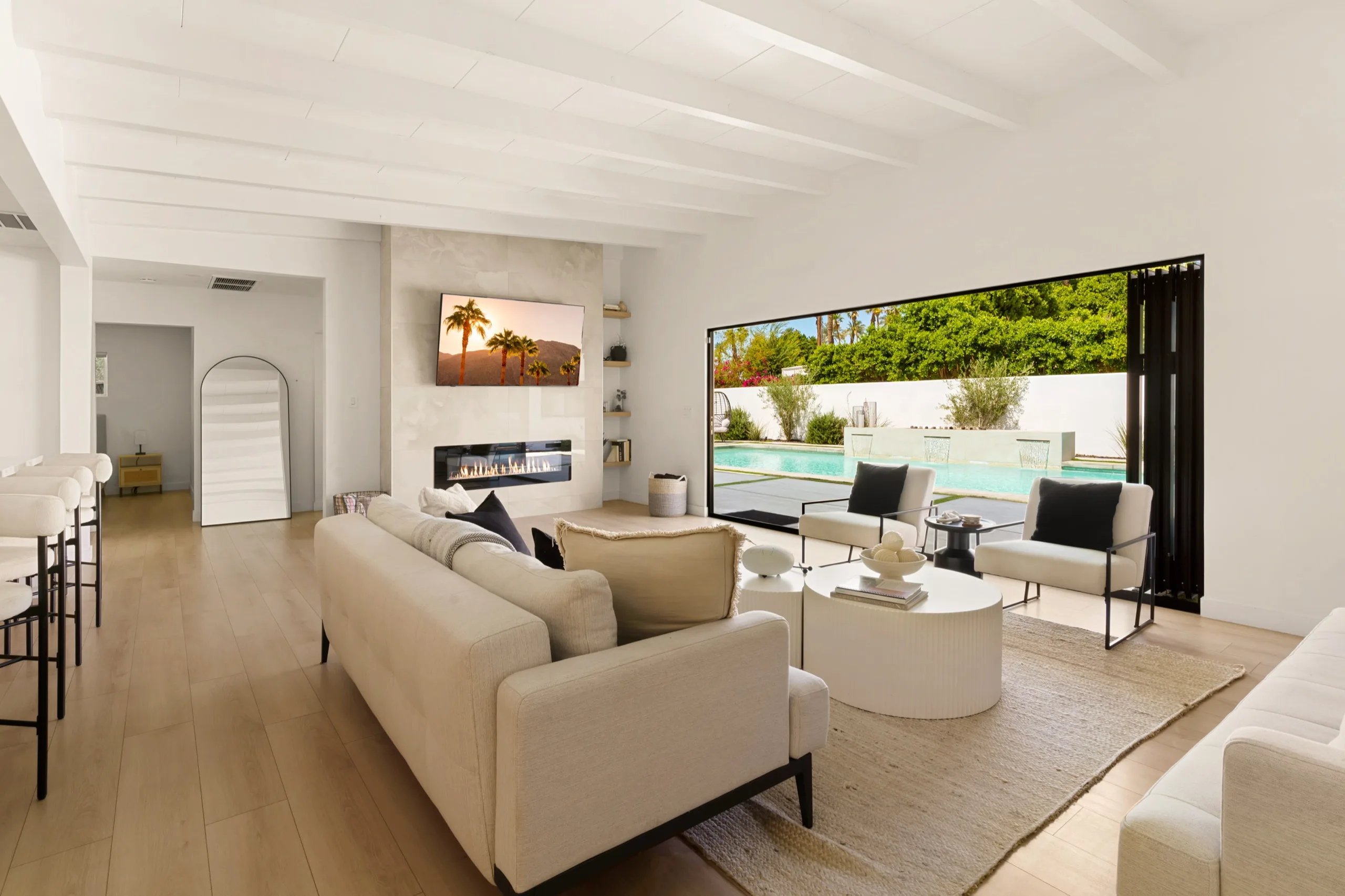 Open-plan living room with pale oak floors and a linen sectional, sliding glass doors folded back to a pool terrace