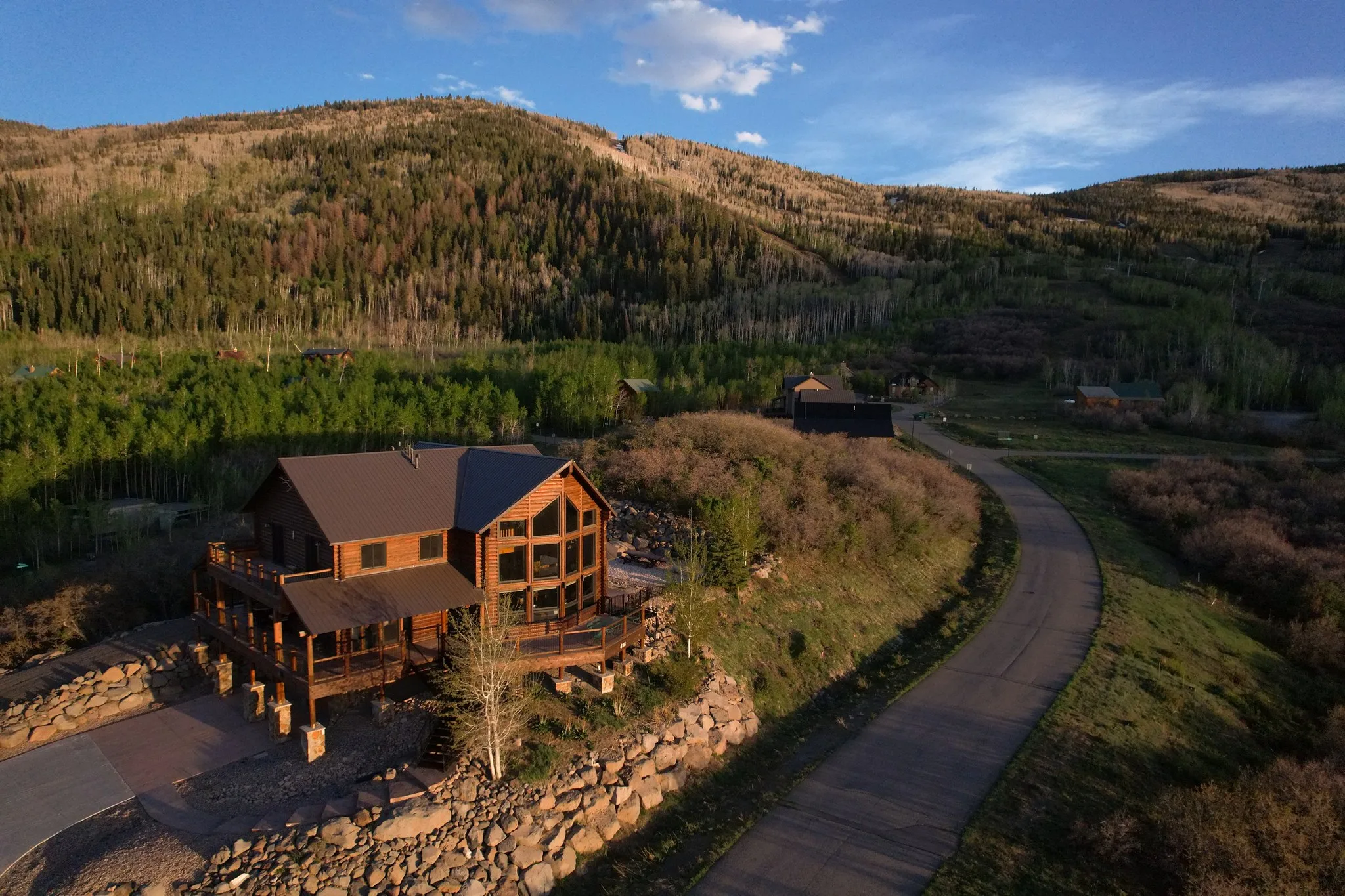 Aerial view of a log house on a hillside above a curving drive, forested ridges rising behind it