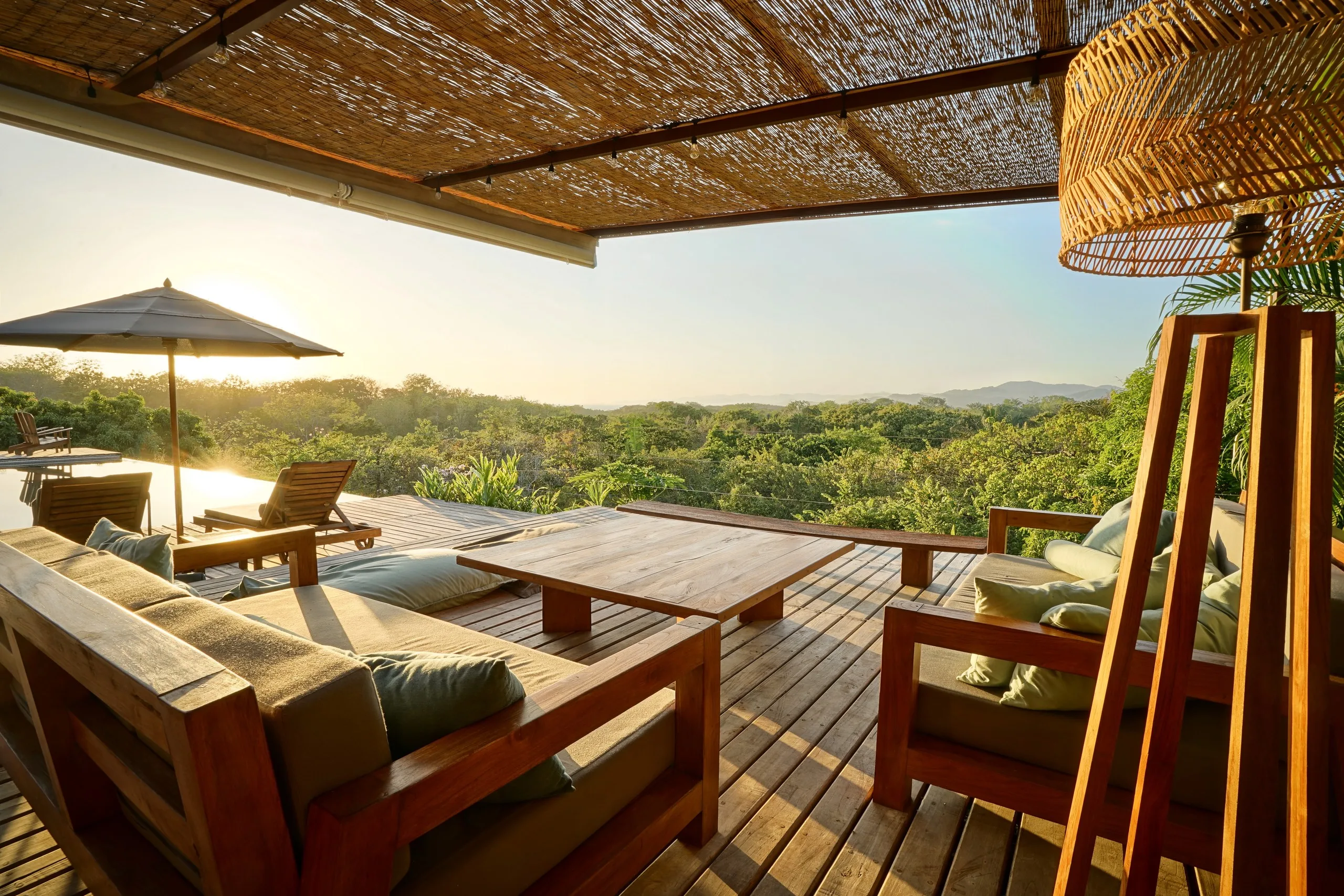 Thatched terrace at golden hour, teak daybeds and a low table above an unbroken canopy of jungle